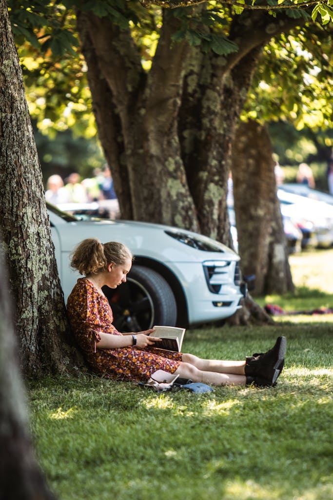 Woman sitting under a tree in a park reading a book near a parked car.